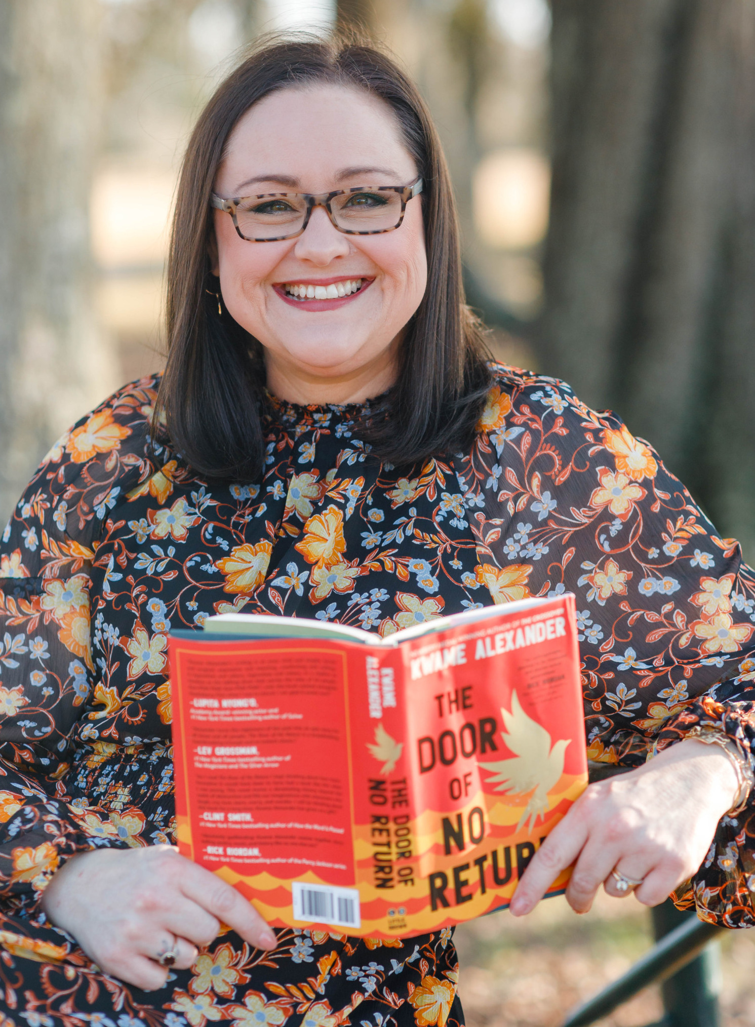 Book bans: Dark-haired woman with glasses in dark floral top smiles into camera while holding a book open with its red cover facing the camera