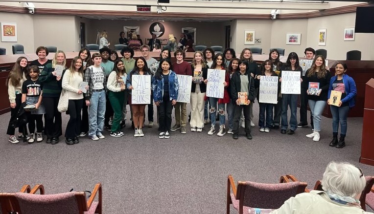 Book bans: Large group of young students stand closely together in large room facing camera holding handmade protest signs.