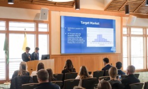 College Male Enrollment: Two young men in navy suits stand at front of seated group next to a presentation slide om bright blue with white text