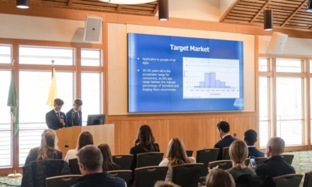 College Male Enrollment: Two young men in navy suits stand at front of seated group next to a presentation slide om bright blue with white text