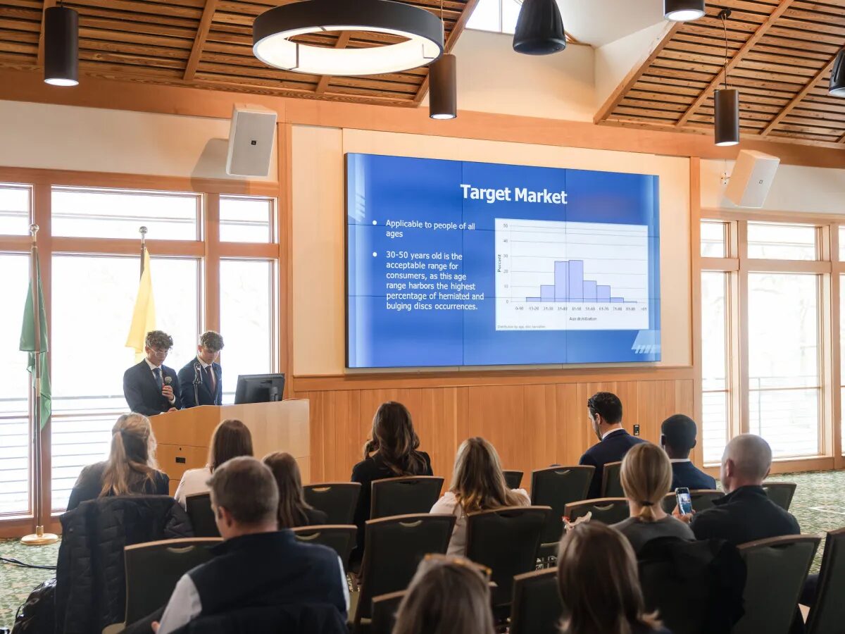 College Male Enrollment: Two young men in navy suits stand at front of seated group next to a presentation slide om bright blue with white text