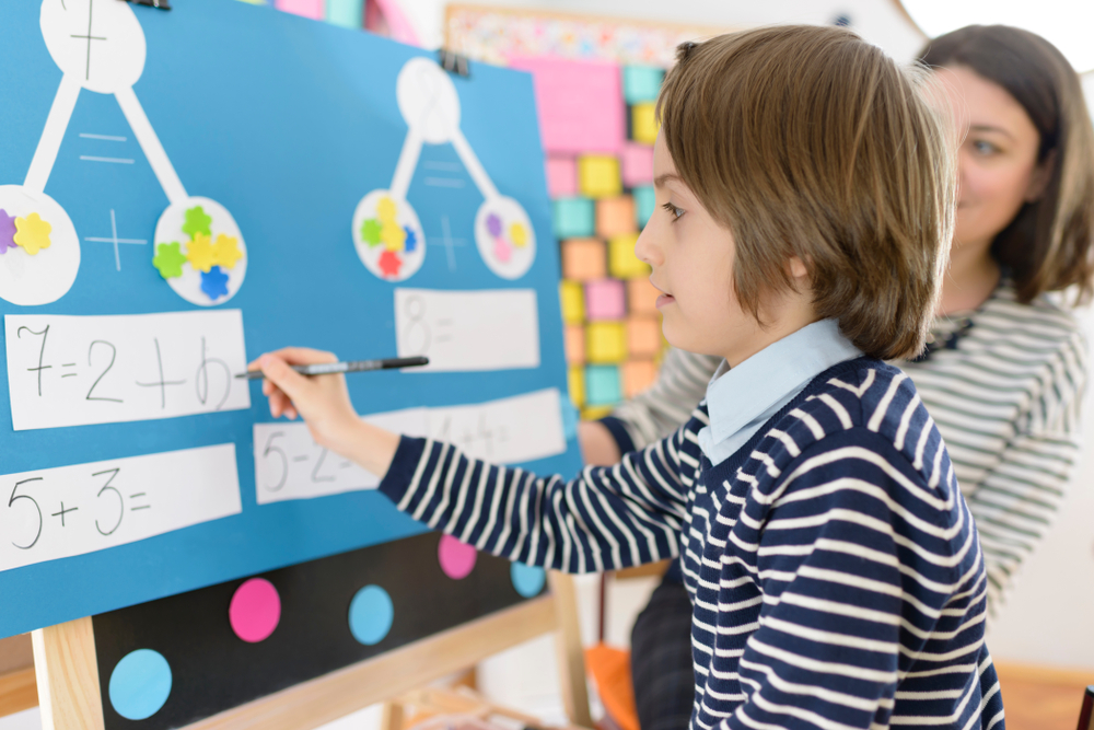 KIndergarten Math: Young boy stands at b;ie paper-covered corkboard doing math problems set up in connected white paper circles