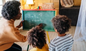 KIndergarten Math: Black woman kneels at small blackboard writing numbers with chalk as two young black children sitting close to board watch