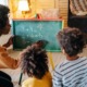 KIndergarten Math: Black woman kneels at small blackboard writing numbers with chalk as two young black children sitting close to board watch