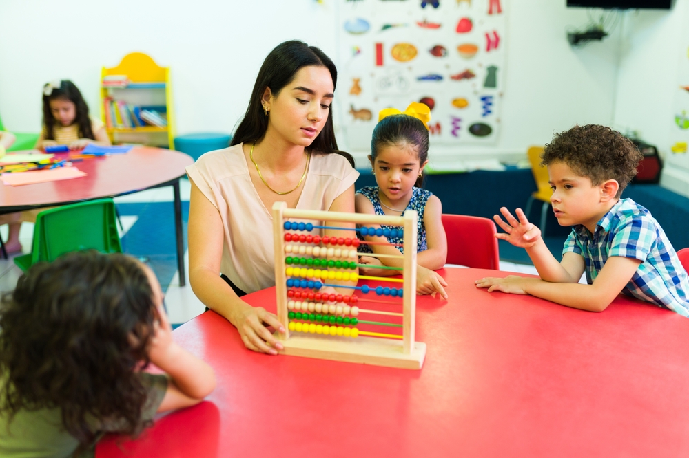 KIndergarten Math: Female teacher sits at table with 4 young students learning to count using a colorful beads-on-straight-wires type abacus.