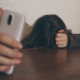 Survey teen depression and smart phones: Person with long dark hair sits with head down on dark brown table top hiding face and holding silver smart phone in right hand with arm on table reaching towards camera