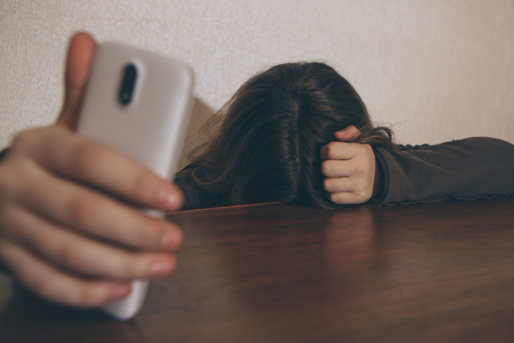 Survey teen depression and smart phones: Person with long dark hair sits with head down on dark brown table top hiding face and holding silver smart phone in right hand with arm on table reaching towards camera