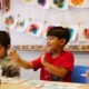 Colorado public preschool: Three preschool bots all with dark fair sit at a long table in front of a colorful bulletin board water coloring