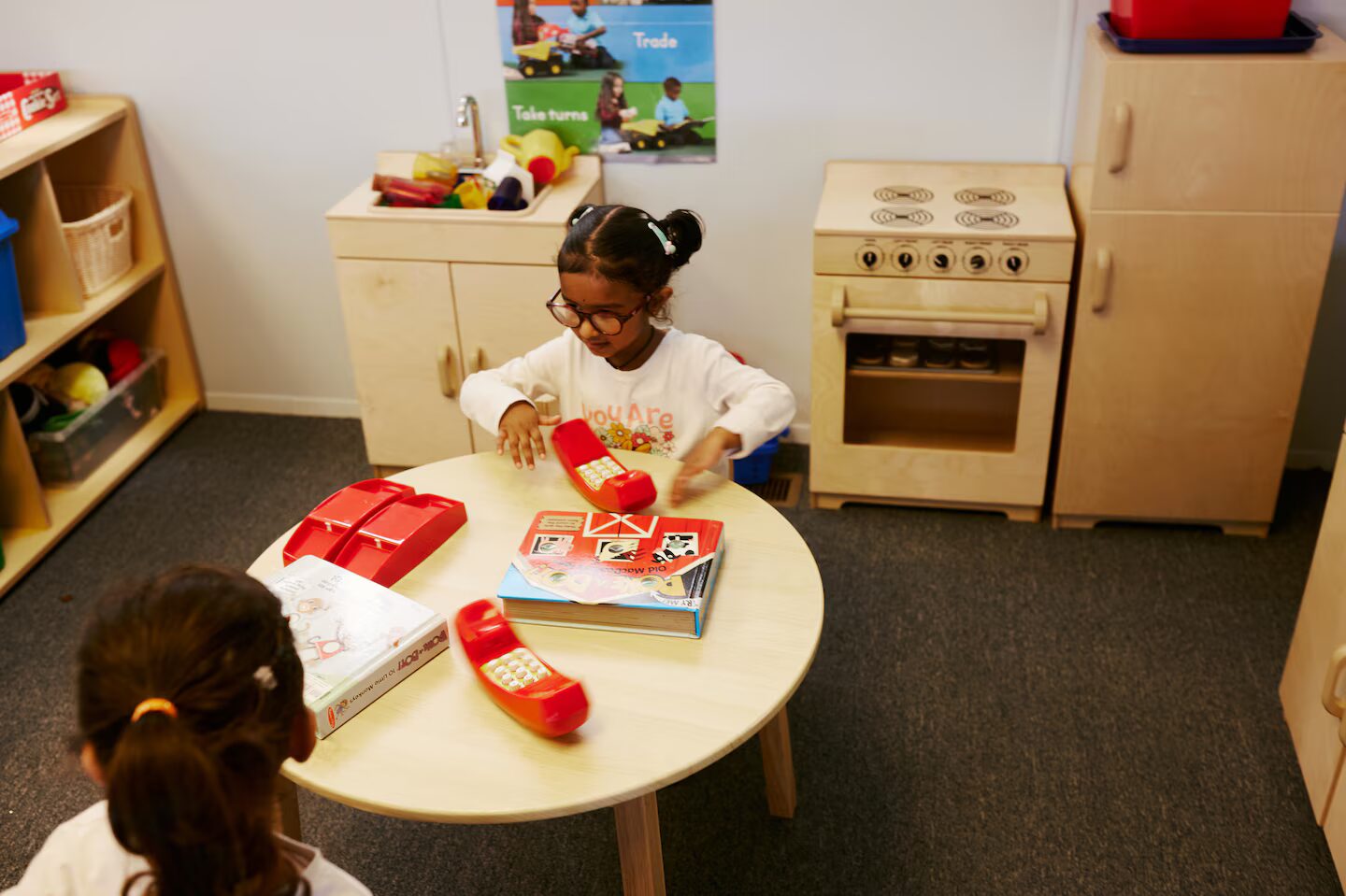 Colorado public preschool: Two preschool girls sit at a low, round table playing with large, red, plastic phones with a play kitchen sink, stove and refrigerator (all in light wood) behind them.