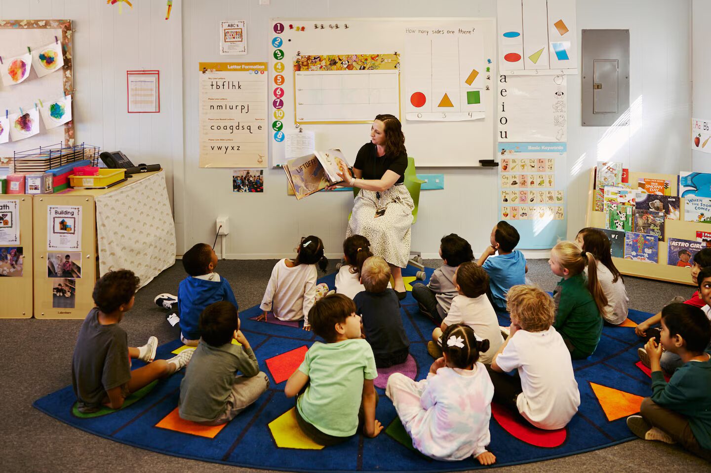Colorado public preschool: Ten preschool children sit on colorful matts on floor of a classroom, in front of a seated, dark-haired, teacher reading a book to them and showing them the book's pictures