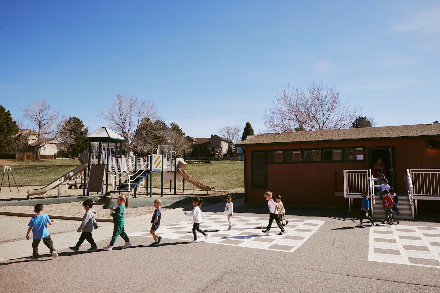 Colorado public preschool: Several preschool children exiting a brown bungalow classroom building going down steps, then walking across an asphalt area with a playground gym on the background.