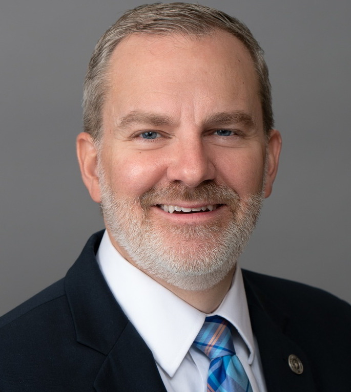 Chaplains in schools: Headshot man with graying blonde hair and gray beard in dark suit with white shirt and blue plaid tie.