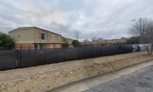 HUD housing lawsuit: Long shot of large, 2-story, apartment complex with many boarded up windows surrounded by dead grass and black, spiked aluminum fence covered in black shade cloth with large grafitti