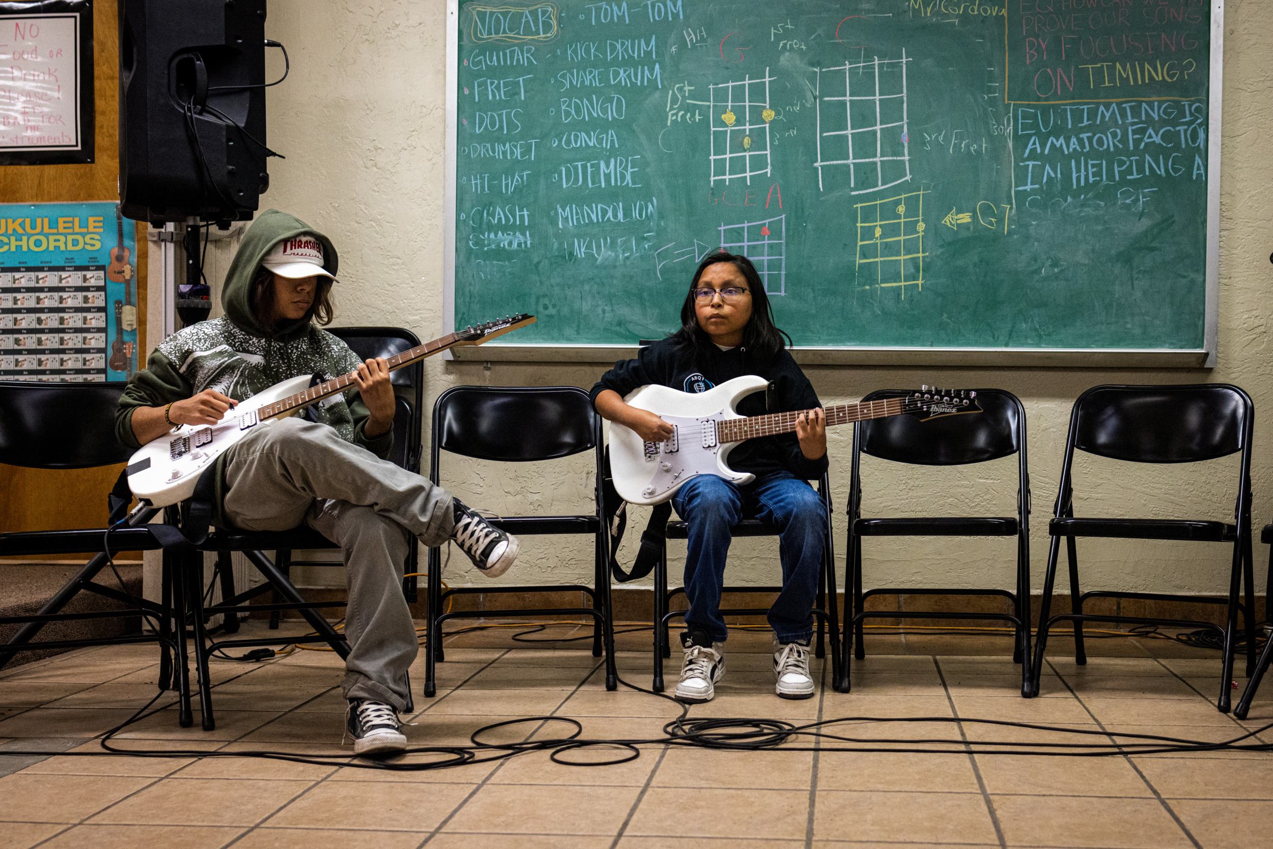 Native American Charter School; Two male teens with dark hair sit in front of a chalkboard and plat guitars