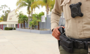 Police on campus: closeup of only middle and right side body showing hand on gun in holster of police on tan and brown uniform with modern high school campus in the background