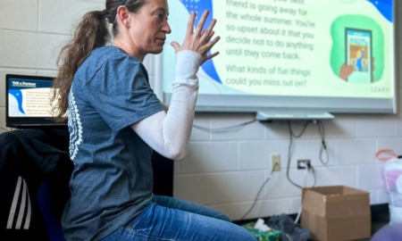 Elementary suspension: Woman with long, loght brown hair pulled back in ponytail in blue top and jeans sits in front of classroom video screen gesturing with both hands in air