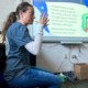 Elementary suspension: Woman with long, loght brown hair pulled back in ponytail in blue top and jeans sits in front of classroom video screen gesturing with both hands in air