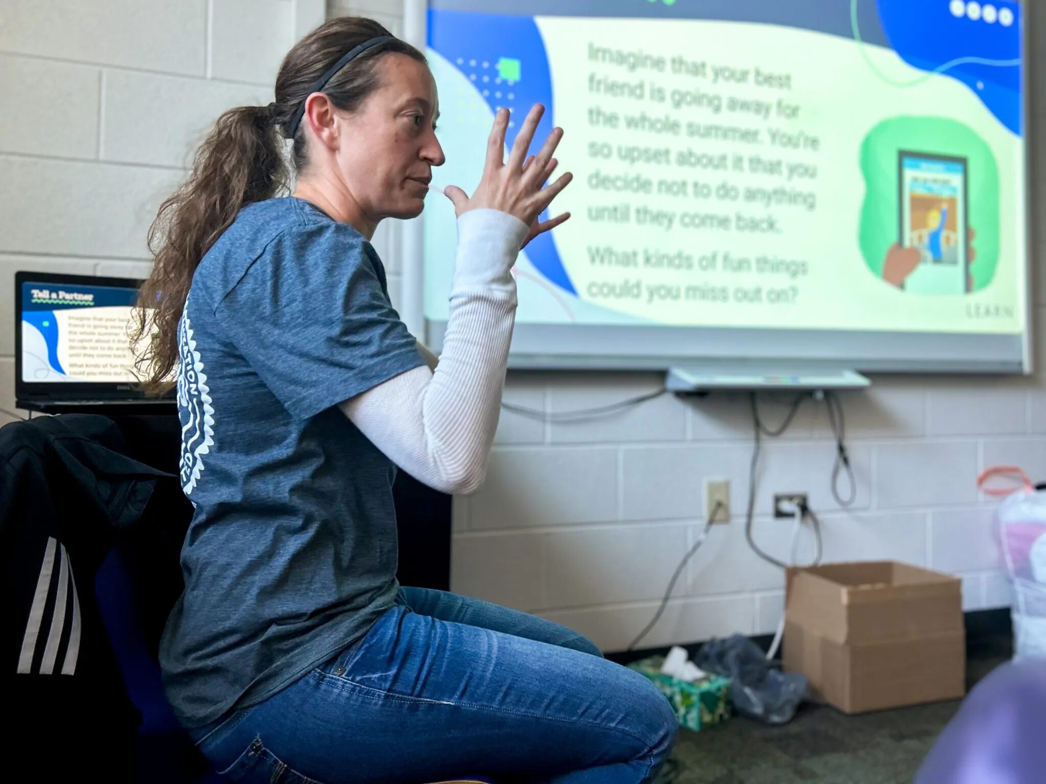 Elementary suspension: Woman with long, loght brown hair pulled back in ponytail in blue top and jeans sits in front of classroom video screen gesturing with both hands in air