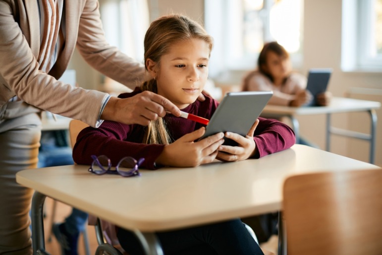 Literacy and touch: Young student in dark red shirt with light brown hair in a long braids sits at school desk holding a tablet looking at screen while teacher next to them points to something on the screen