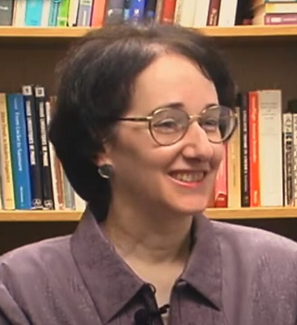 Literacy and touch: Headshot of smiling woman with gold-rimmed glasses and short brunette hair in brown, collared shirt, sitting in front of fill bookshelves