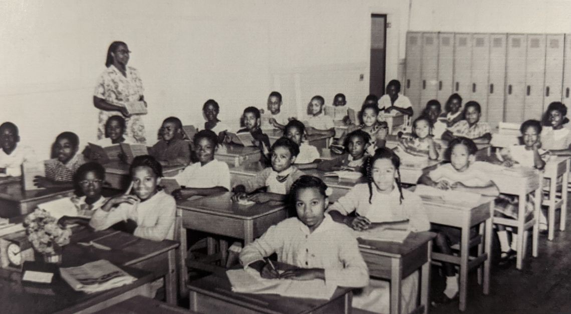 Classroom of all Black students seated in 4 rows of desks with female Black woman teacher standing against wall to left of desks