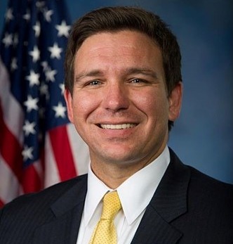Academic freedom: Man with dark brown hair on black suit, white shirt and gold tie with part of USA flag in background