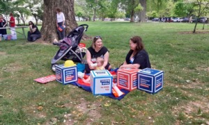 Parents tote toddlers to D.C. for child tax credit and child care: two women, one with a baby, sitting and talking on a blanket in a park with protest materials