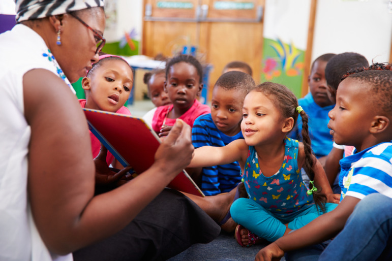 Pre-school skills research: Black female teacher reads a book to several Black pre-school students seated in the floor.