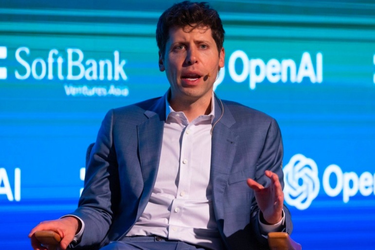 Teachers & AI usage: Sam Altman in blue/grey suit jacket and white shirt sits on conference stage on front of teal and turquoise backdrop speaking into small headset microphone.