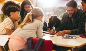 Teacher layoffs: Group of elementary school kids stand around a table in a classroom, attentively leaning over table and following a lesson being taught by their male teacher with glasses, dark hair and beard.