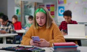 School cell phone bans: Teenage schoolgirl with green hair chatting on smartphone sitting at desk in class. Teen student using cellphone in classroom at school