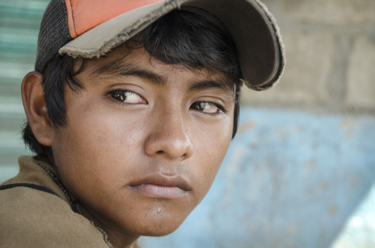 Older Immigrant student school admissions: Close up of wistftul face of a male Latino teen with dark hair wearing a faded orange and ran baseball cap.