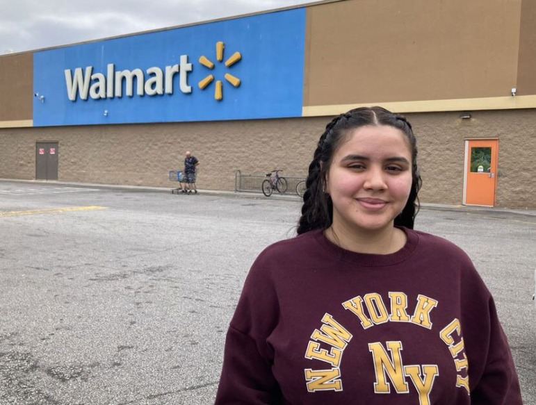 Immigrant student school admissions: Young Latino woman with long dark hair on maroon sweatshirt stands in front of a tan building with Walmart signage