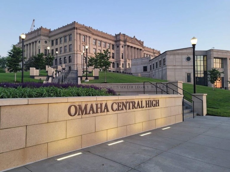 Immigrant student school admissions: Long front view up a sloping, grass lawn of two, tan stone and brick multi-story buildings - one modern, one traditional - with tan stone wall signage on sidewalk reading Omaha Central High