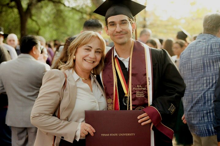 Immigrant student school admissions: Latino woman with long blonde hair in beige suit and white blouse stands next to white young man with dark hair in black college graduation gown and cap both holding a side of a brown covered college diploma