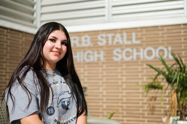 Older Immigrant student school admissions: Smiling Latinx girl with very long black hair in gray r-shirt standing in front of a red brick wall with name o high school on ot