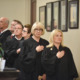 Oklahoma-strikes-down-religious-charter-school: Several people in black judge's robes stand at attention on interior steps with hand over heart facing American flag