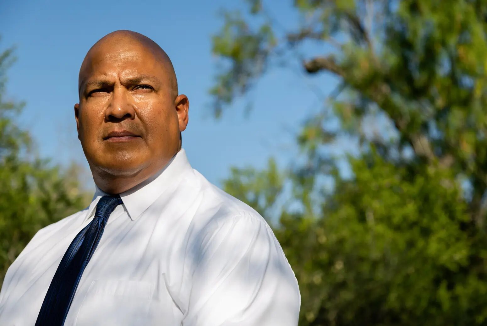 Uvalde indictment: Bald white man in white business shirt and dark tie stands staring seriously into camera under blue sky with green trees in the background.