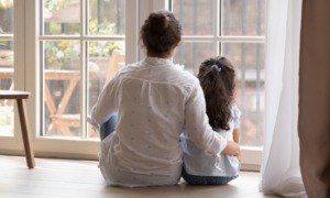 child sexual abuse prevention: back view of dark-haired woman in white shirt with arm round the back of young, dark-haired child, both seated in front of large window looking outside