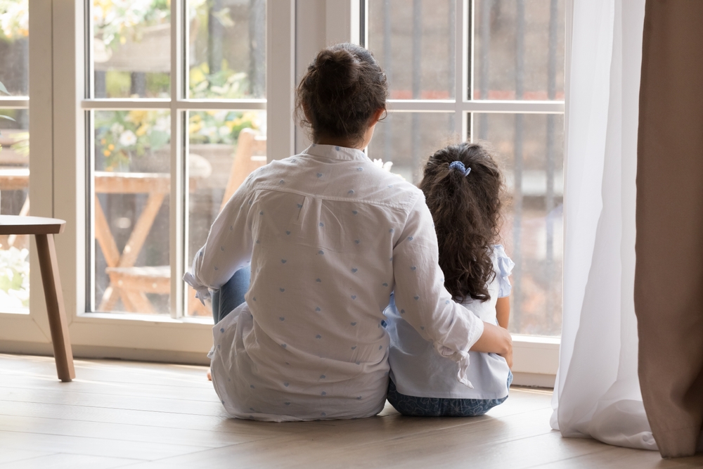 child sexual abuse prevention: back view of dark-haired woman in white shirt with arm round the back of young, dark-haired child, both seated in front of large window looking outside
