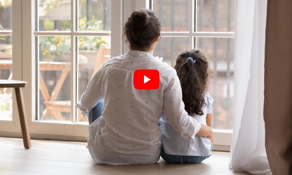 child maltreatment and abuse prevention: back view of dark-haired woman in white shirt with arm round the back of young, dark-haired child, both seated in front of large window looking outside