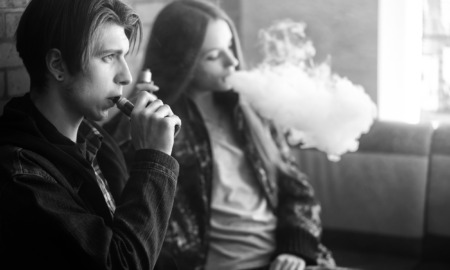 Colorado Vaping Lawsuit settlement: Black and white photo of two older teens sitting against brick wall smoking electronic cigarettes