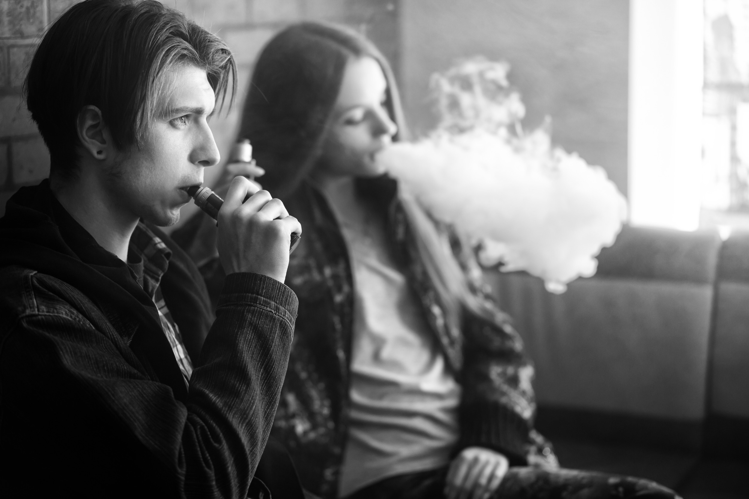 Colorado Vaping Lawsuit settlement: Black and white photo of two older teens sitting against brick wall smoking electronic cigarettes