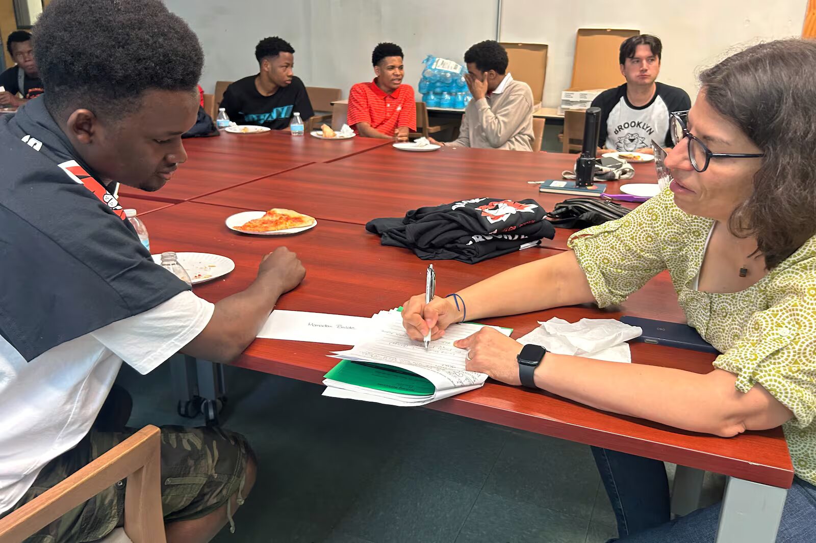 Older Immigrant students high school enrollment: Group of Black, male, older teens sit stound wood conference table with older brown-haired white woman helping one in foreground to fill out paperwork