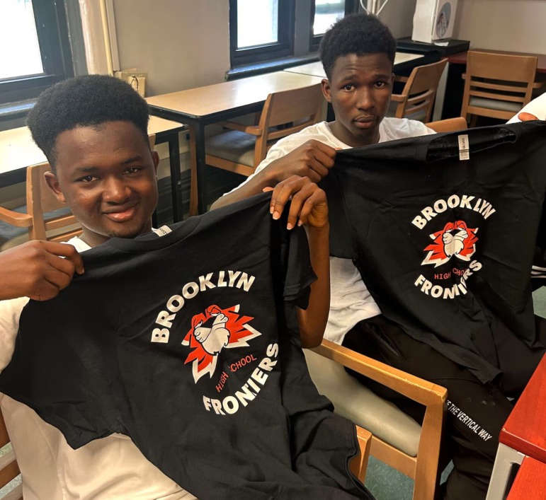 Older Immigrant students high school enrollment: Two older black male teens sit in chairs holding up black t-shirts with red and white logo and text "Brooklynn Frontiers."
