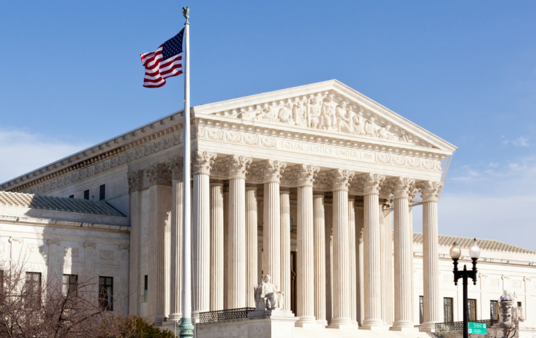 Bible teaching Oklahoma public schools: Facade of US Supreme court — a large, multi-story, white, traditional architecture structure with many pillars and a waving US flag on pole yo left side of entrance steps — in Washington DC on sunny day.