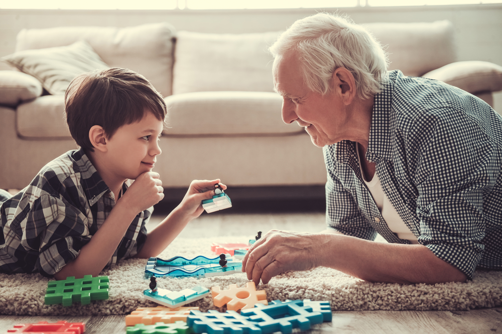 Kinship foster care: Older white-haired man and young boy are playing with toys on the carpeted floor, looking at each other and smiling
