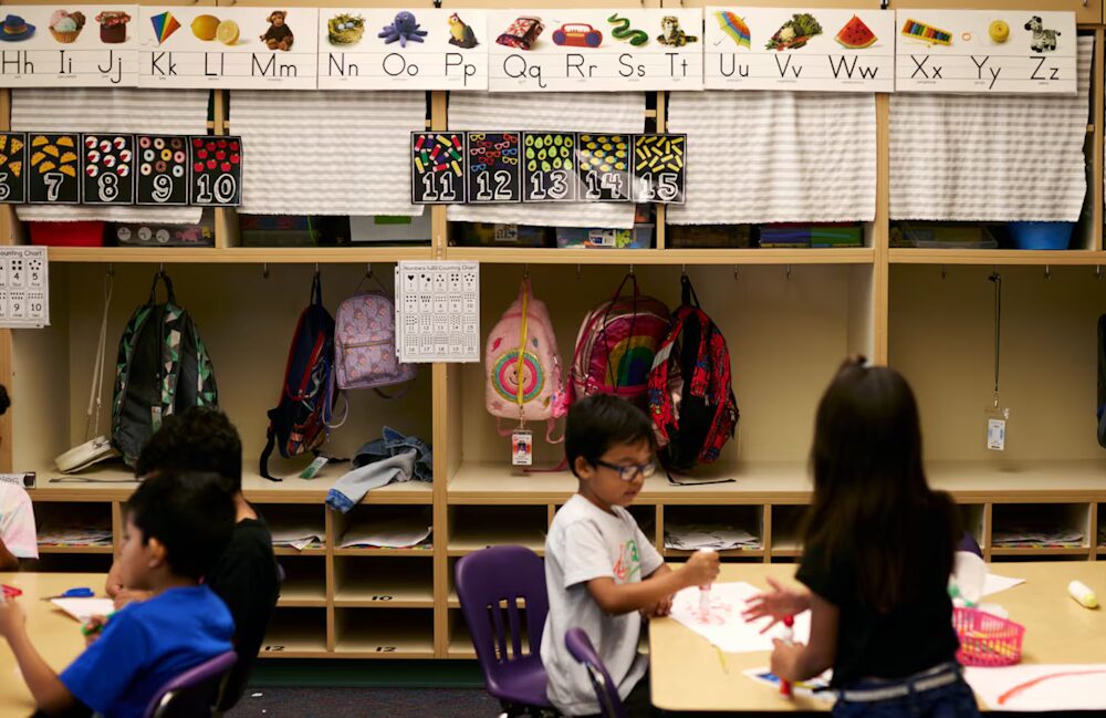 Undocumented students: Three young, dark-haired students sit at low tables in elementary classroom in front of wooden cubbies and colorful alphabet wall decorations