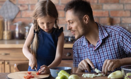 Math talk by parents to children: Adult man with short brown hair in blue plaid shirt talks to young girl with long blonde hair in navy dress as both stand at kitchen counter chopping vegetables