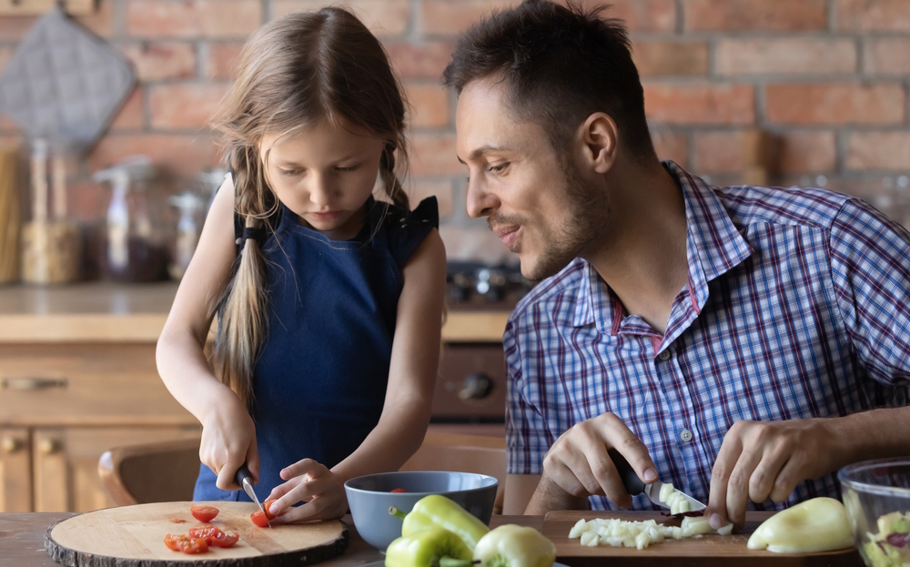 Math talk by parents to children: Adult man with short brown hair in blue plaid shirt talks to young girl with long blonde hair in navy dress as both stand at kitchen counter chopping vegetables